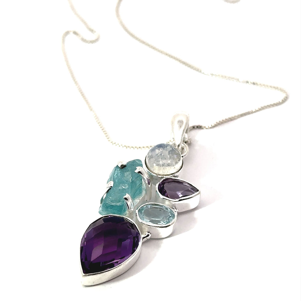 The Amethyst Multi Stone Pendant is a silver necklace featuring an amethyst pendant adorned with a captivating blend of purple, blue, and turquoise gemstones set against a white background, ideal for lovers of bohemian jewelry.