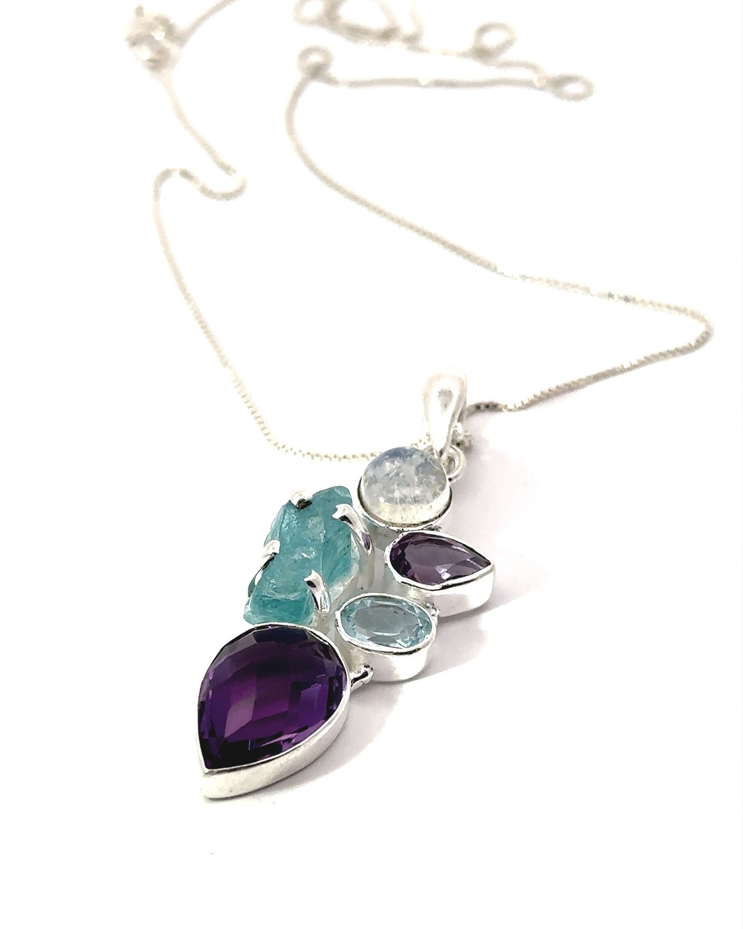 The Amethyst Multi Stone Pendant is a silver necklace featuring an amethyst pendant adorned with a captivating blend of purple, blue, and turquoise gemstones set against a white background, ideal for lovers of bohemian jewelry.