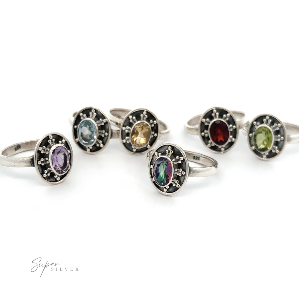Six Oval Gemstone Rings with Ball and Disk Border in different colors (purple, blue, yellow, red, green) set in circular black bases are arranged in two rows. The words 