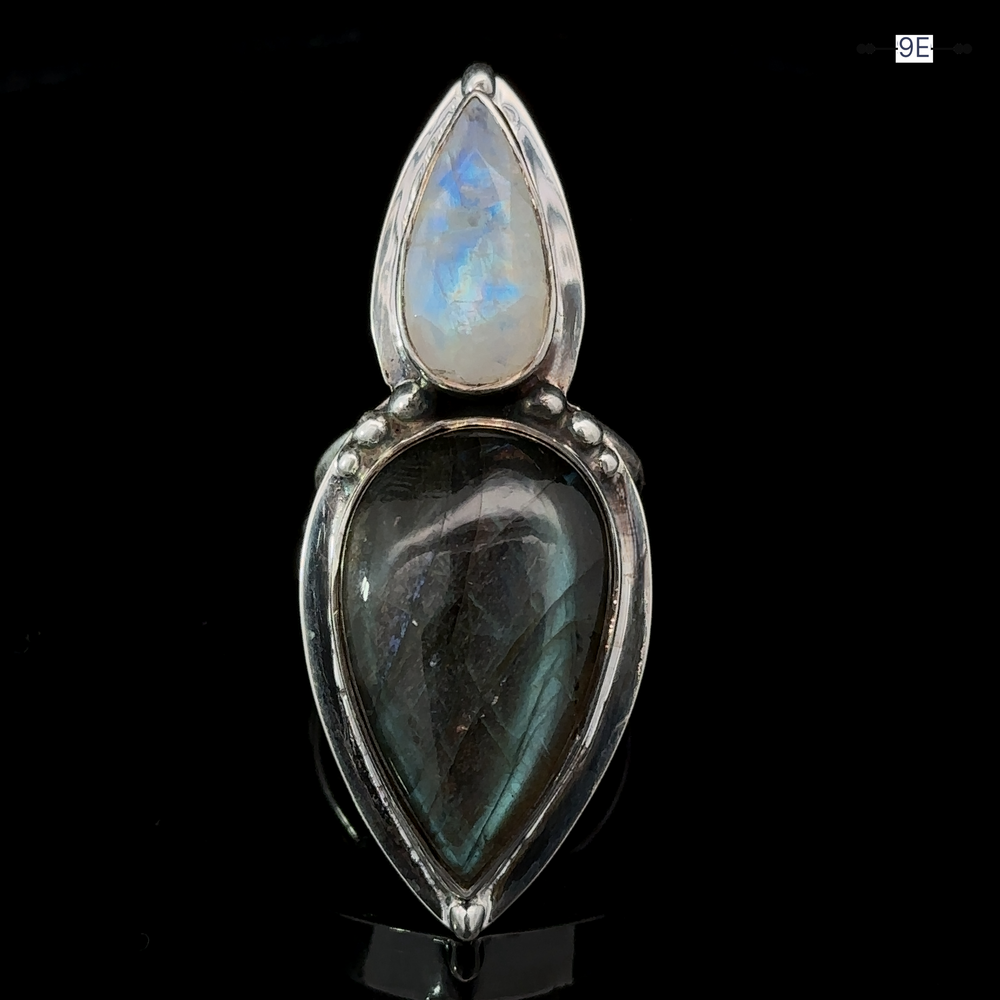 
                  
                    The Heavy Labradorite And Moonstone Ring is a silver statement piece featuring two tear-shaped gemstones; the upper stone is a moonstone with a translucent blue sheen, while the lower stone is labradorite with a darker greyish hue, set against a black background.
                  
                