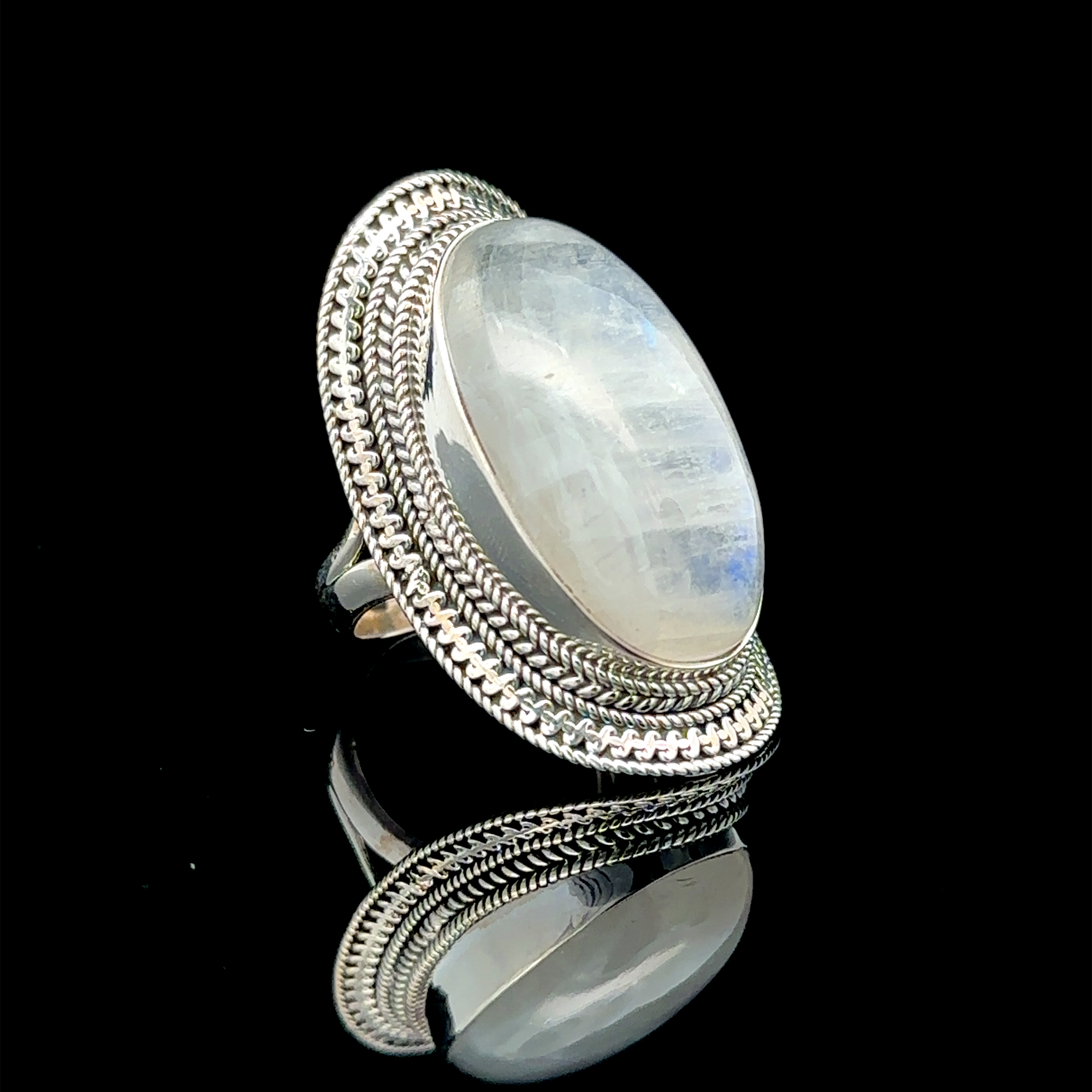 A Large Oval Shield Gemstone Ring with a large, oval, milky white gemstone set within a decorative braided band, displayed against a black background, exuding a subtle bohemian flair.