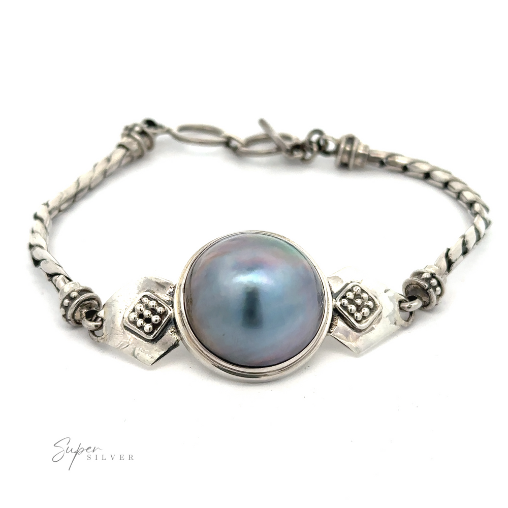 A Blue Mabe Pearl Bali Bracelet with a large central blue Mabe pearl, featuring a braided chain and decorative metal accents. The Bali crafted design is detailed and intricate.