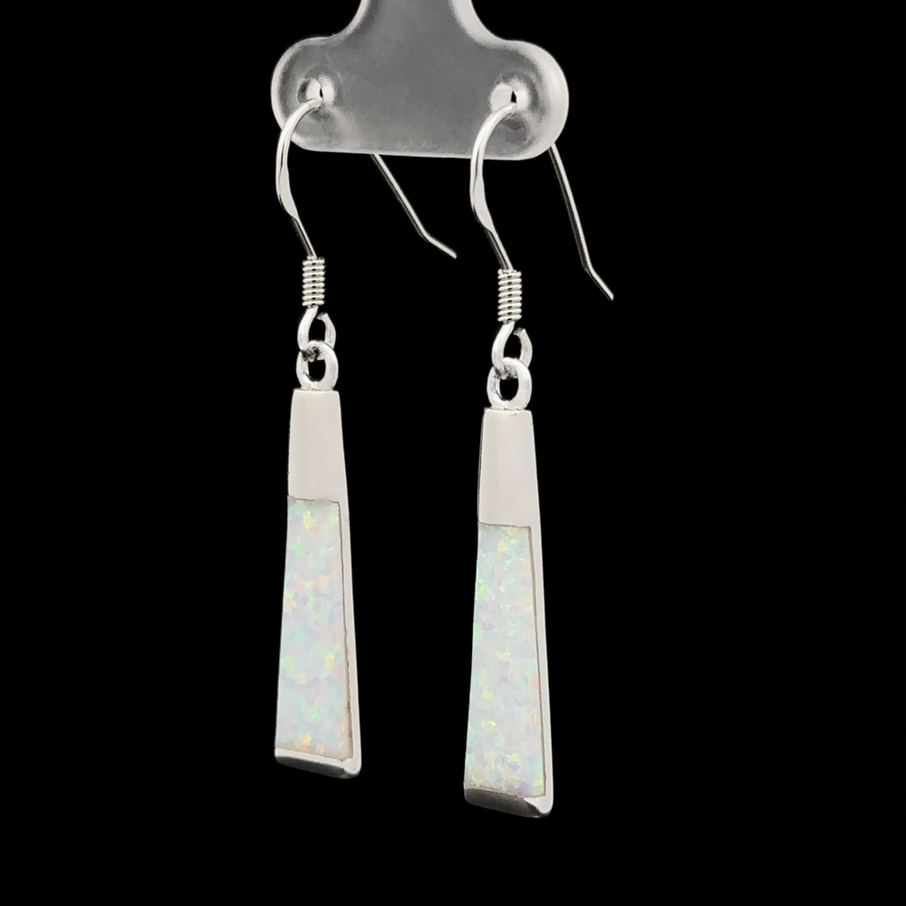 
                  
                    The Elegant Long Opal Earrings feature angular, silver lab-created opals with an iridescent inlay and an elongated flared rectangle shape. Hanging from hooks against a black background, they exude modern elegance.
                  
                