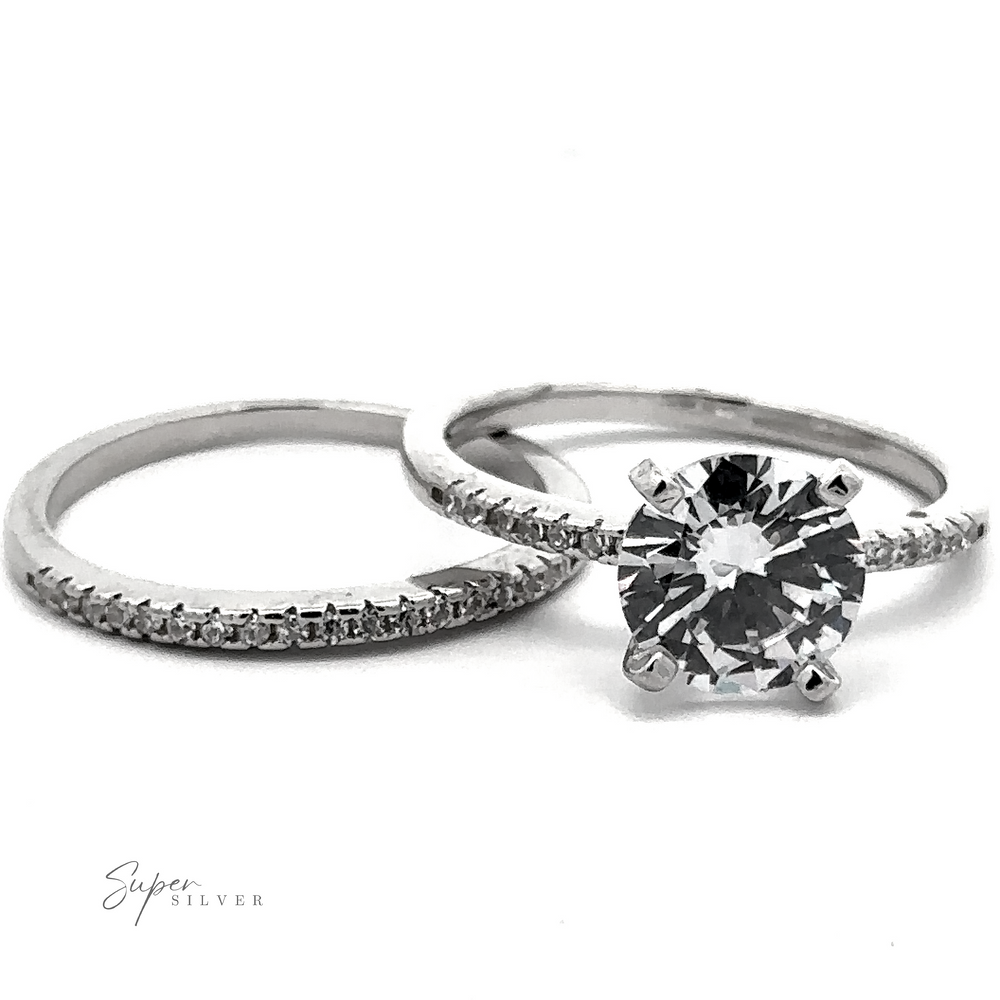 
                  
                    Two silver rings are shown: one is a simple band with small, pave accents, and the other is a round solitaire engagement ring with a large central diamond. "Round Solitaire Wedding Band Set" is inscribed in the corner.
                  
                