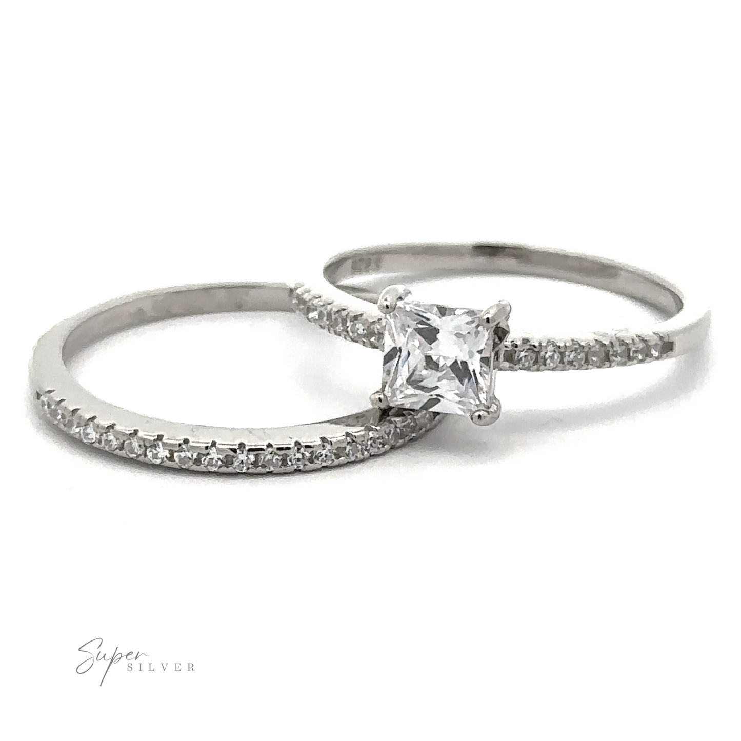 A pair of silver rings with small embedded stones, featuring a rhodium finish. One ring showcases a square-cut central stone, while the other is a simple band adorned with petite gems. This elegant Princess Cut Cubic Zirconia Wedding Band Set perfectly combines sophistication and timeless beauty.