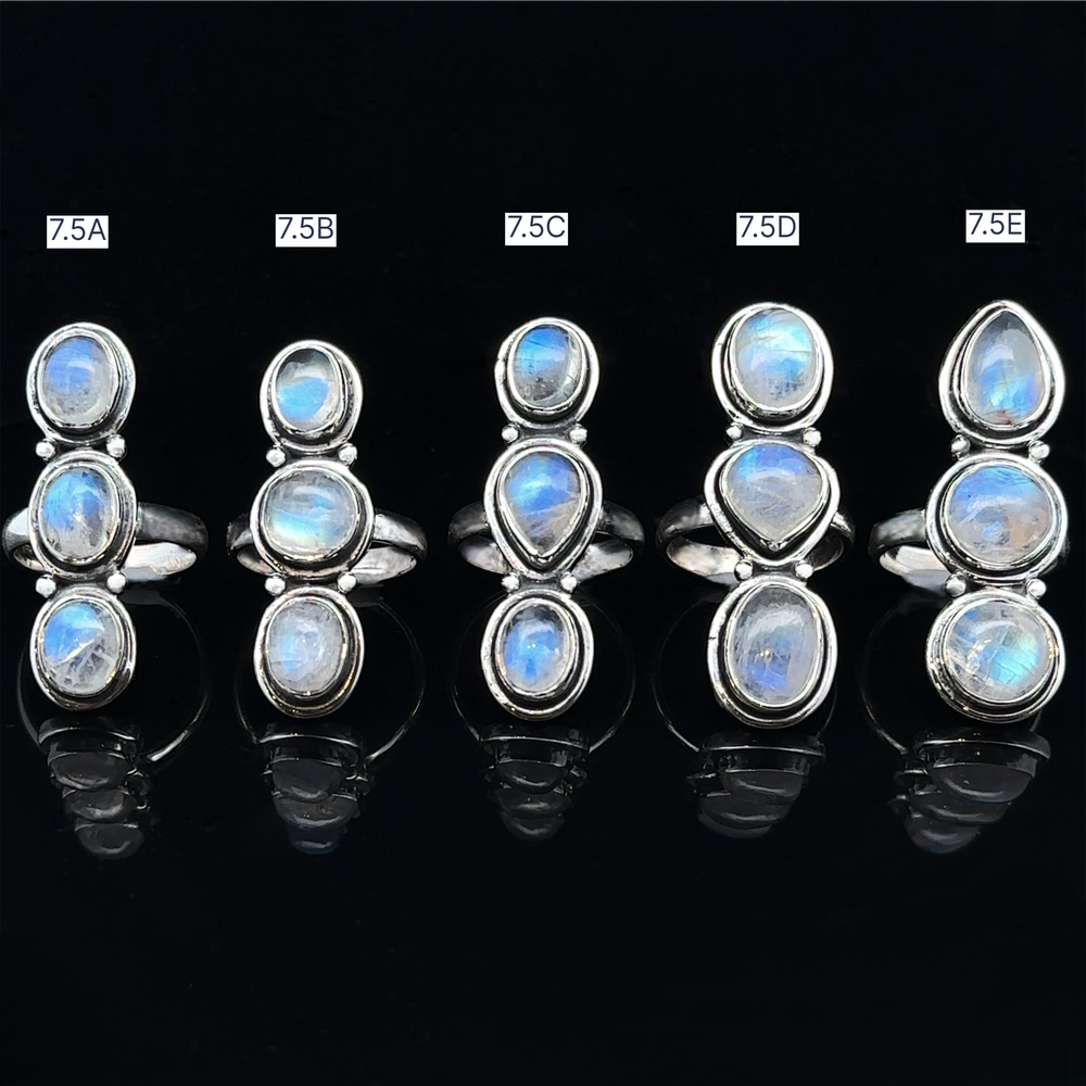 
                  
                    Five Triple Moonstone Rings with blue gemstones, labeled 7.5A to 7.5E, are elegantly displayed on a black background, each emanating a unique goddess energy.
                  
                