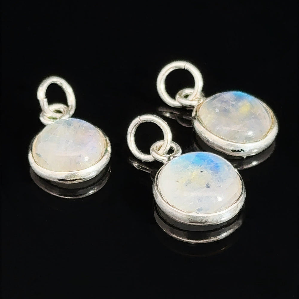 Three Simple and Dainty Moonstone Pendants with sterling silver bezels and loops, placed on a black surface. The shimmering stones catch the light beautifully, each one elegantly wire-wrapped to perfection.