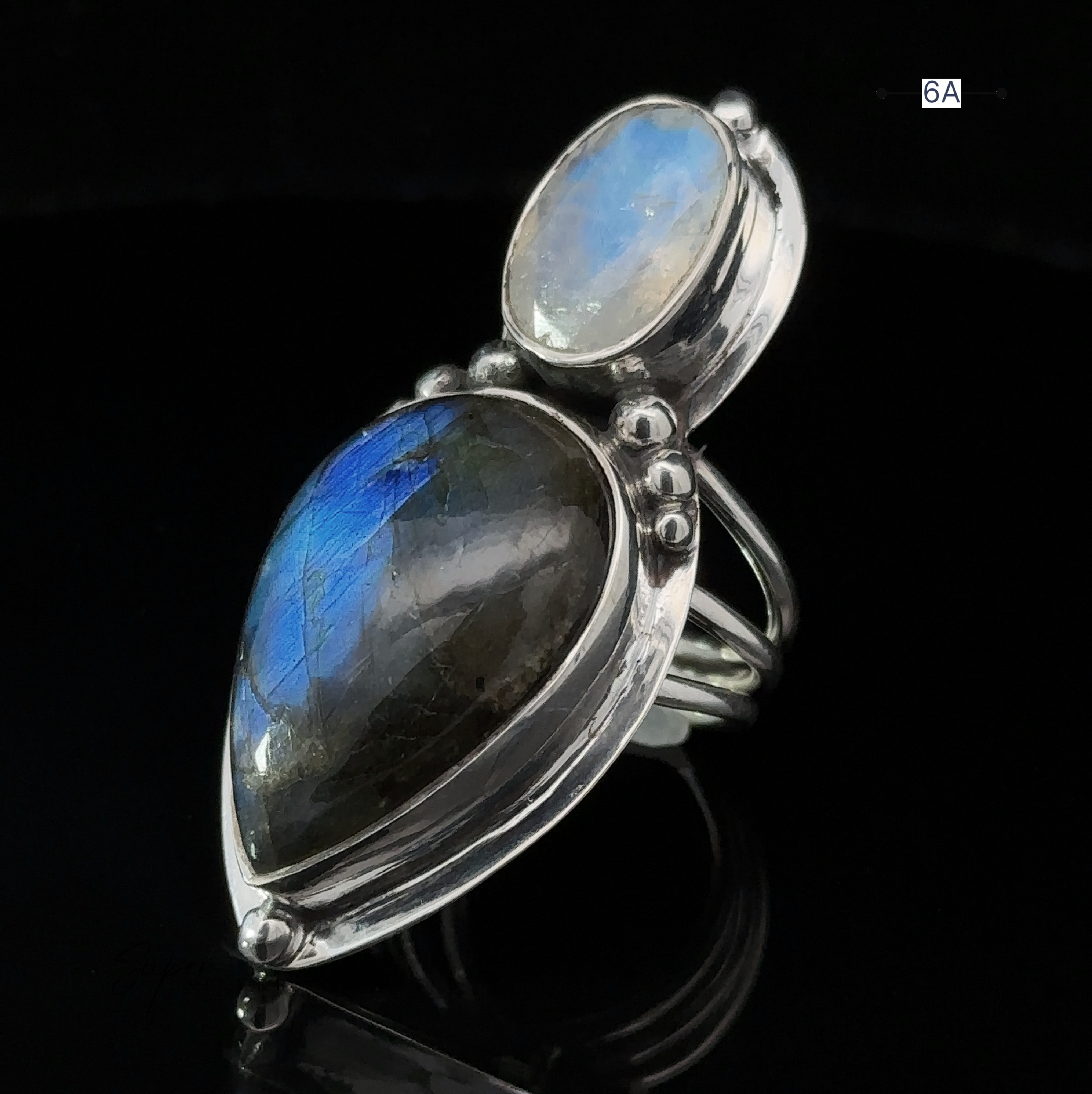 The Heavy Labradorite and Moonstone Ring showcases a striking blue labradorite teardrop stone paired with a smaller oval moonstone, all elegantly set on a dark reflective surface.