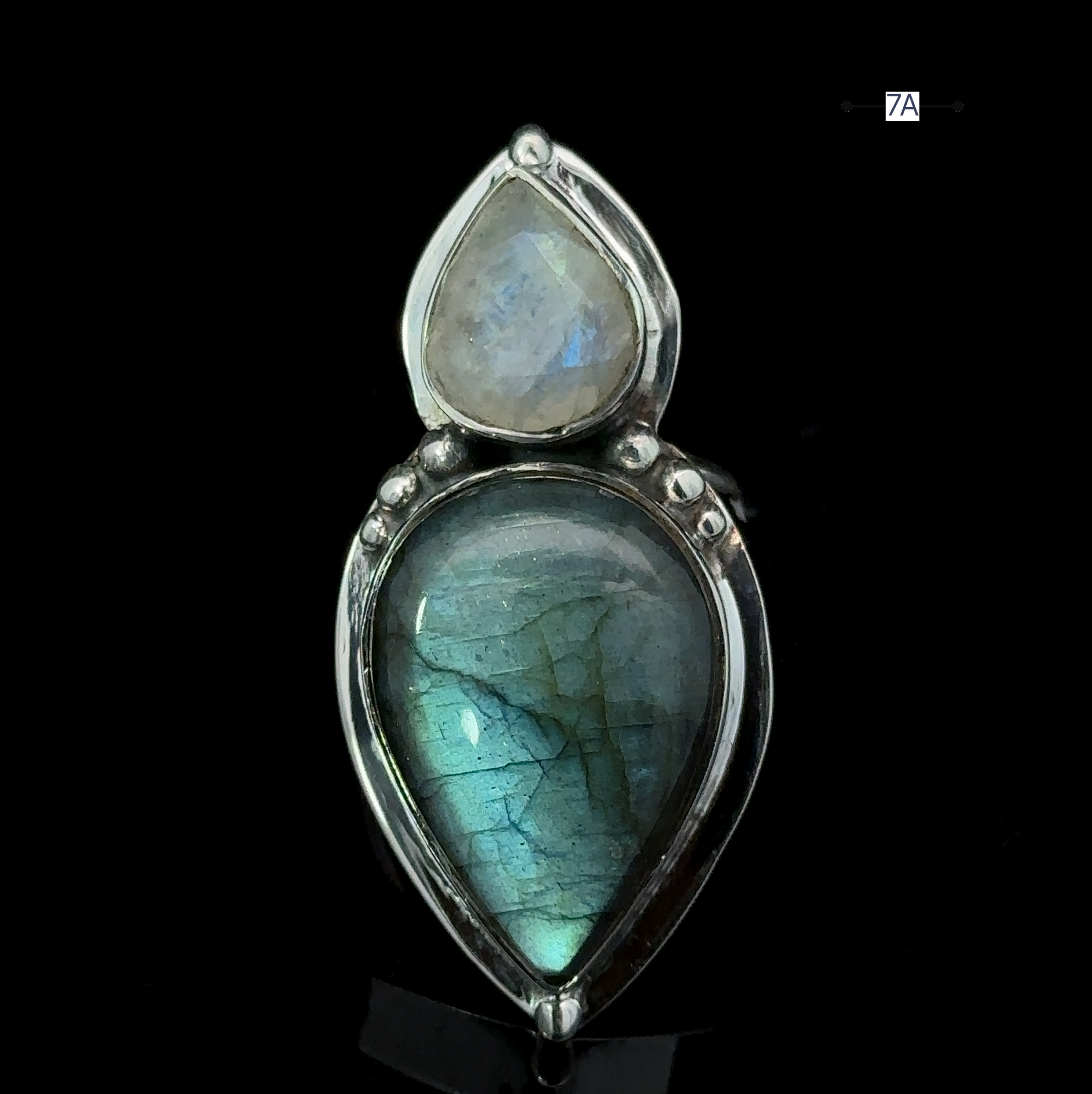 A dual-stone pendant featuring a polished blue labradorite teardrop set in silver below a smaller moonstone is elegantly displayed on a black background, reminiscent of the charm found in the Heavy Labradorite And Moonstone Ring.
