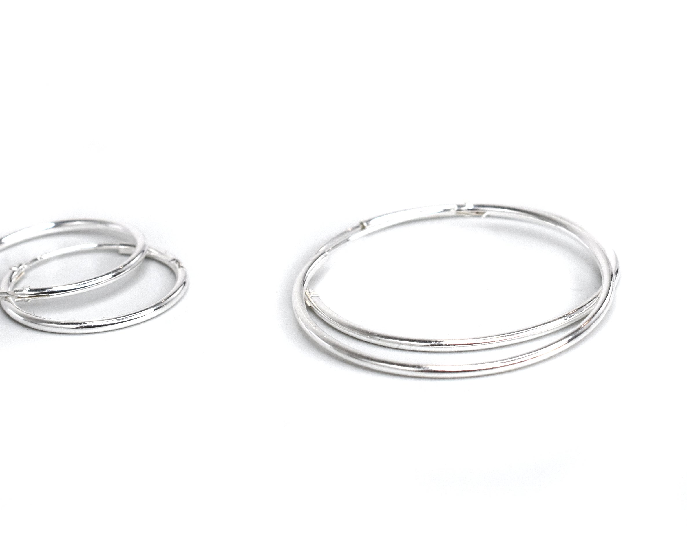 The 1.2mm Infinity Hoops, crafted from .925 Sterling Silver, rest gracefully on a white background with one pair larger than the other. Their subtle elegance and infinity hoop design add a timeless touch to any jewelry collection.