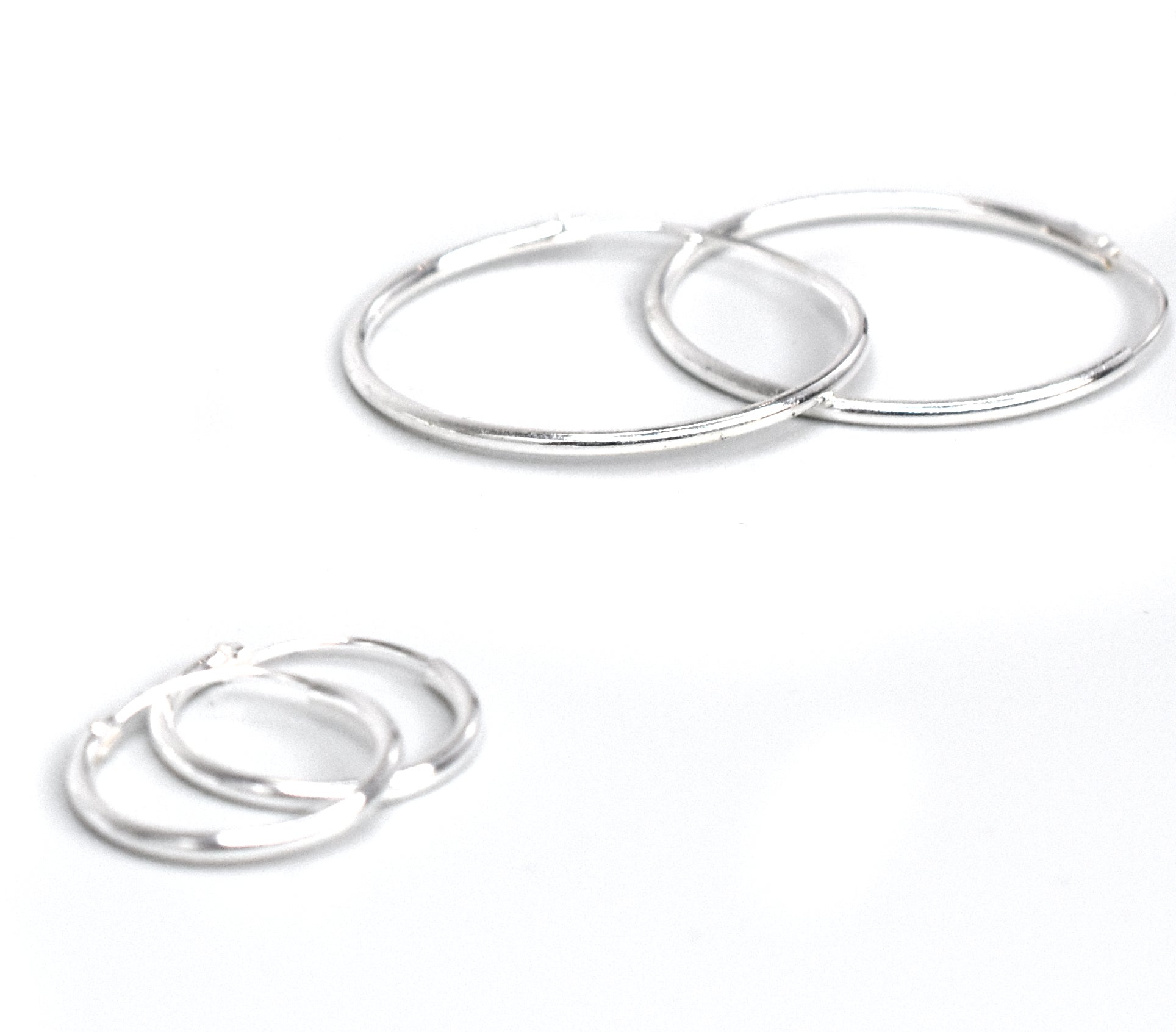 Resting gracefully on a white surface are four elegant earrings crafted from .925 Sterling Silver. Two larger pairs, known as the 1.2mm Infinity Hoops, stand in the back, while two smaller hoops elegantly enhance the front.