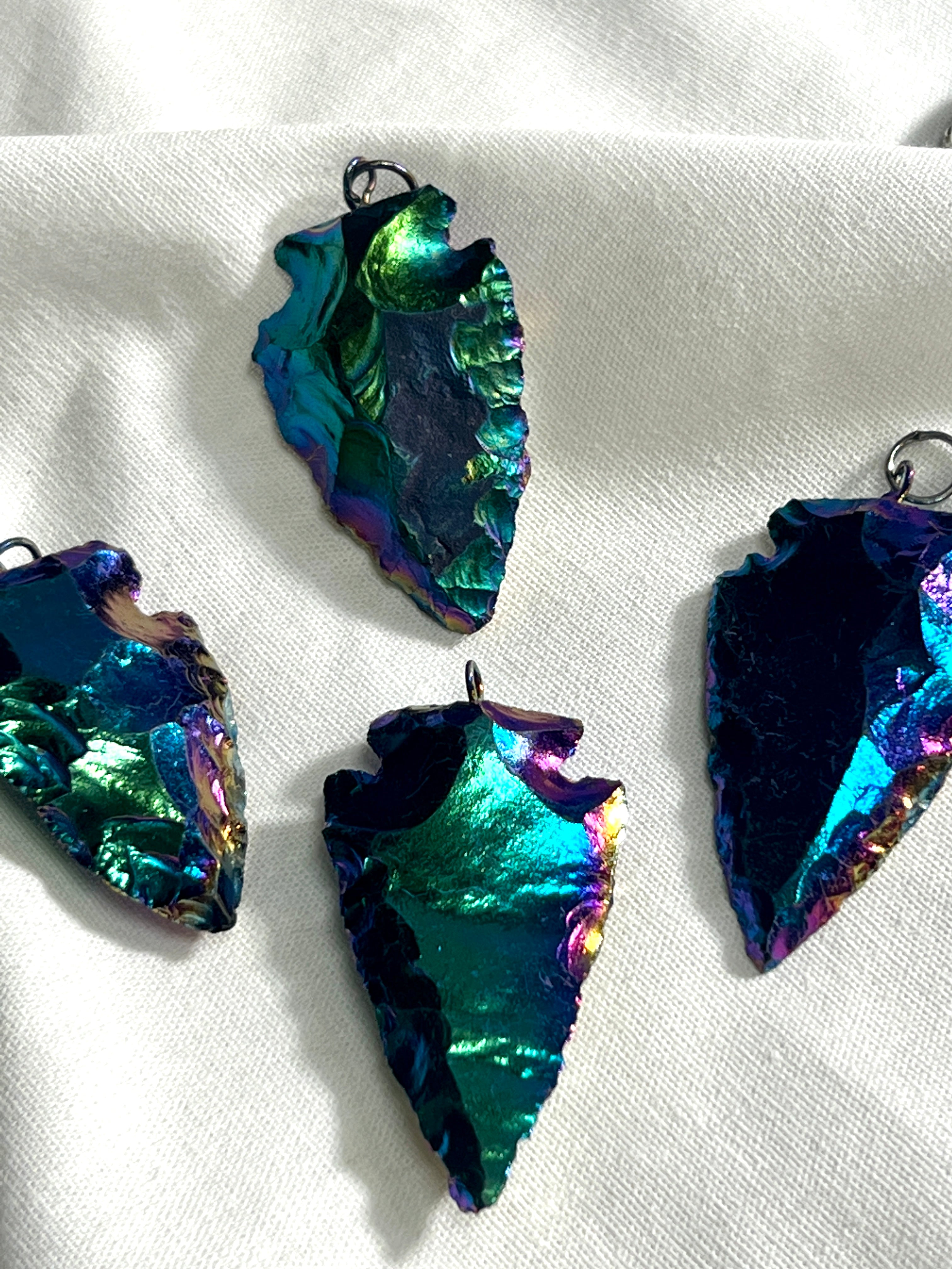 A group of Rainbow Hematite Arrowhead Pendants on a white cloth, suspended from a Super Silver chain.
