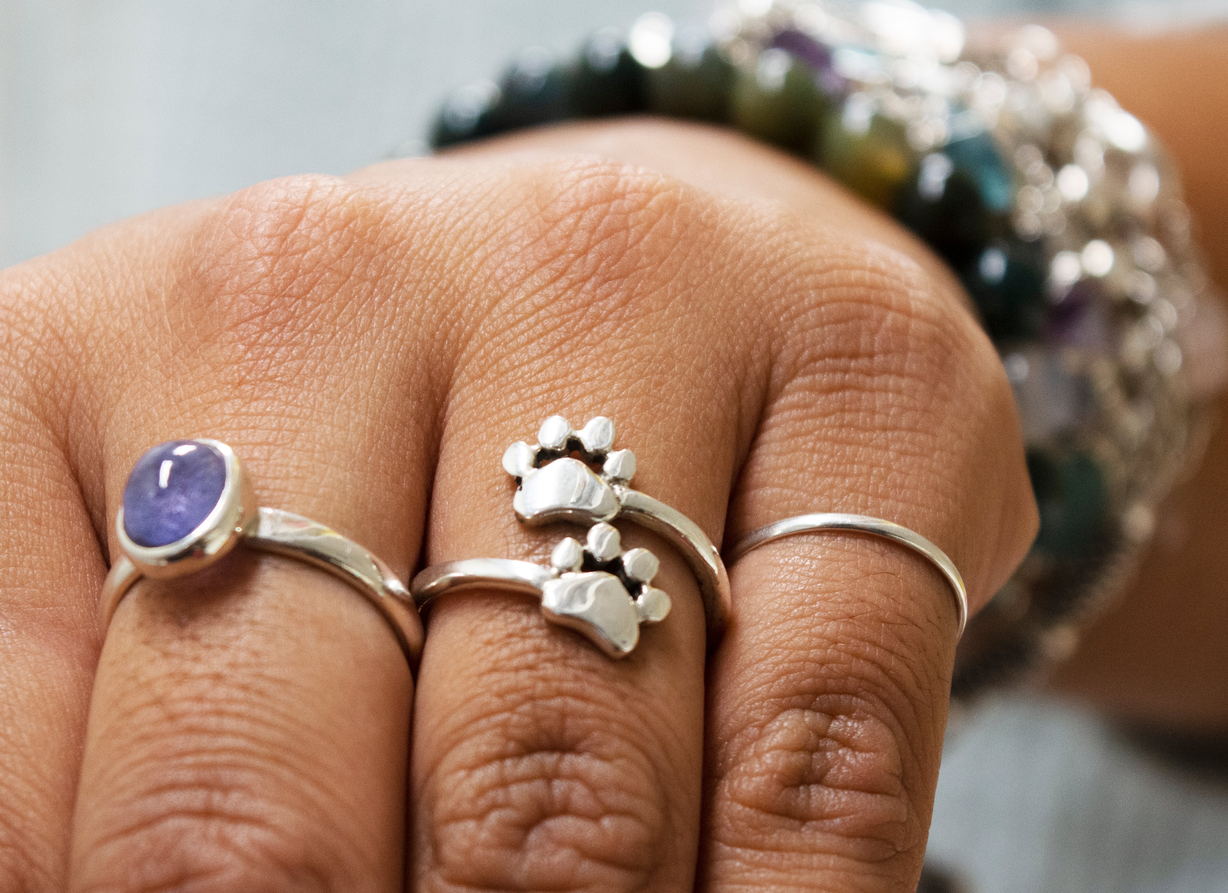 A hand decorated with three silver rings includes the Paw Print Adjustable Ring, ideal for pet lovers, along with a sterling silver ring featuring a purple gemstone. In the background, a multicolored beaded bracelet adds a vibrant touch.