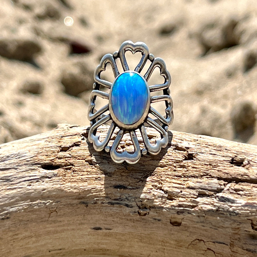 
                  
                    A Super Silver American Made Opal Flower Ring with Heart Shaped Petals, resting on top of a piece of driftwood.
                  
                