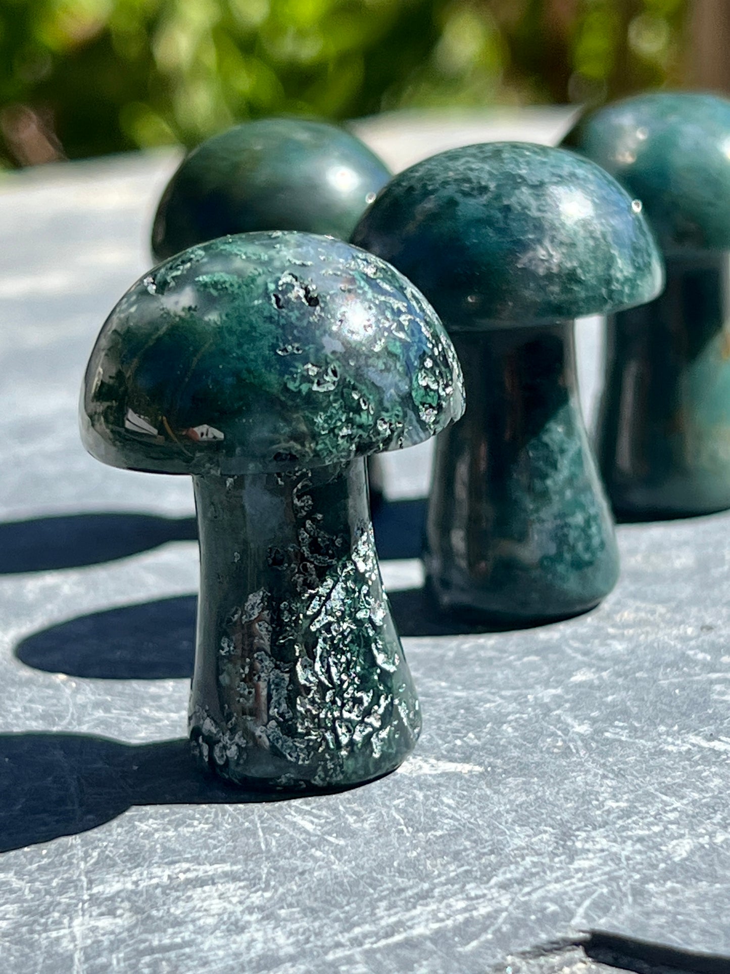 
                  
                    Four Moss Agate Mushrooms sitting on top of a stone table.
                  
                