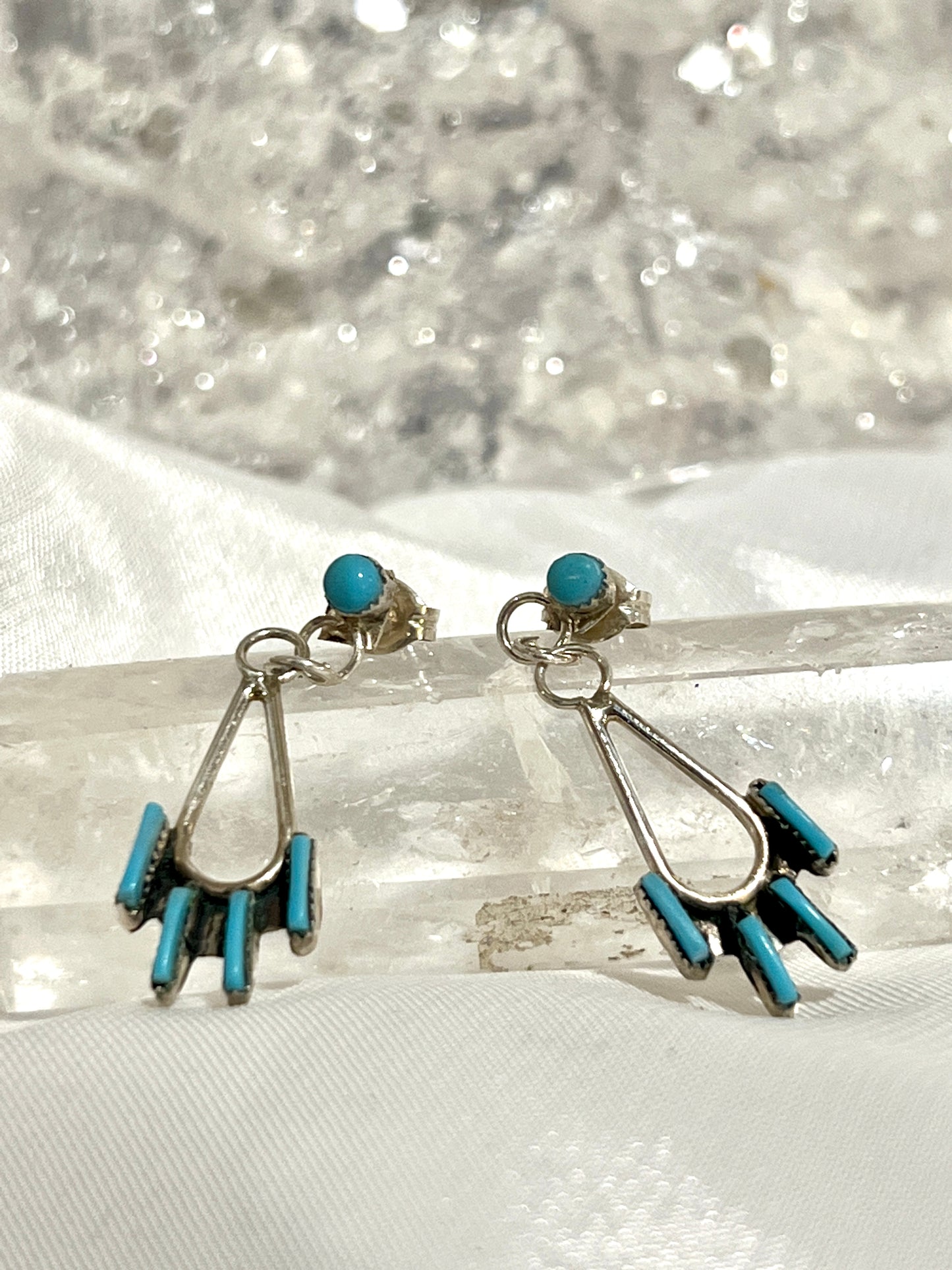 
                  
                    A pair of Super Silver Delicate Needlepoint Zuni Turquoise Earrings sitting on top of a crystal.
                  
                