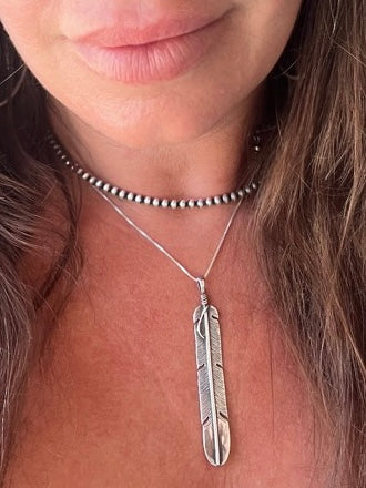 
                  
                    A woman wearing an edgy Super Silver Handcrafted Navajo Pearl Choker adorned with a feather pendant, perfect for creating a rustic southwest collar look.
                  
                