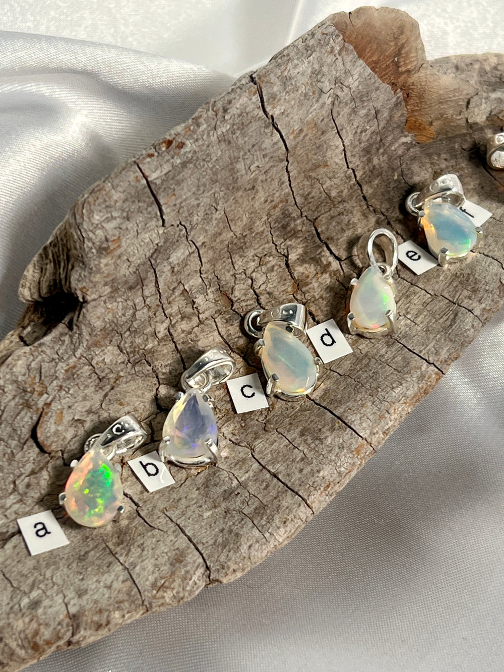 A stunning Dainty Prong Set Facet Cut Teardrop Shaped Ethiopian Opal pendant, delicately crafted with genuine Super Silver settings, suspended on a piece of wood.