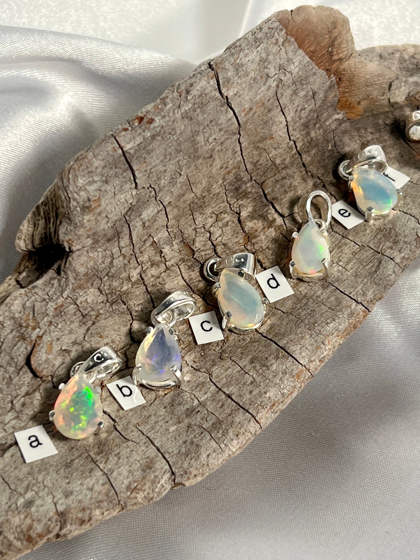 A stunning Dainty Prong Set Facet Cut Teardrop Shaped Ethiopian Opal pendant, delicately crafted with genuine Super Silver settings, suspended on a piece of wood.