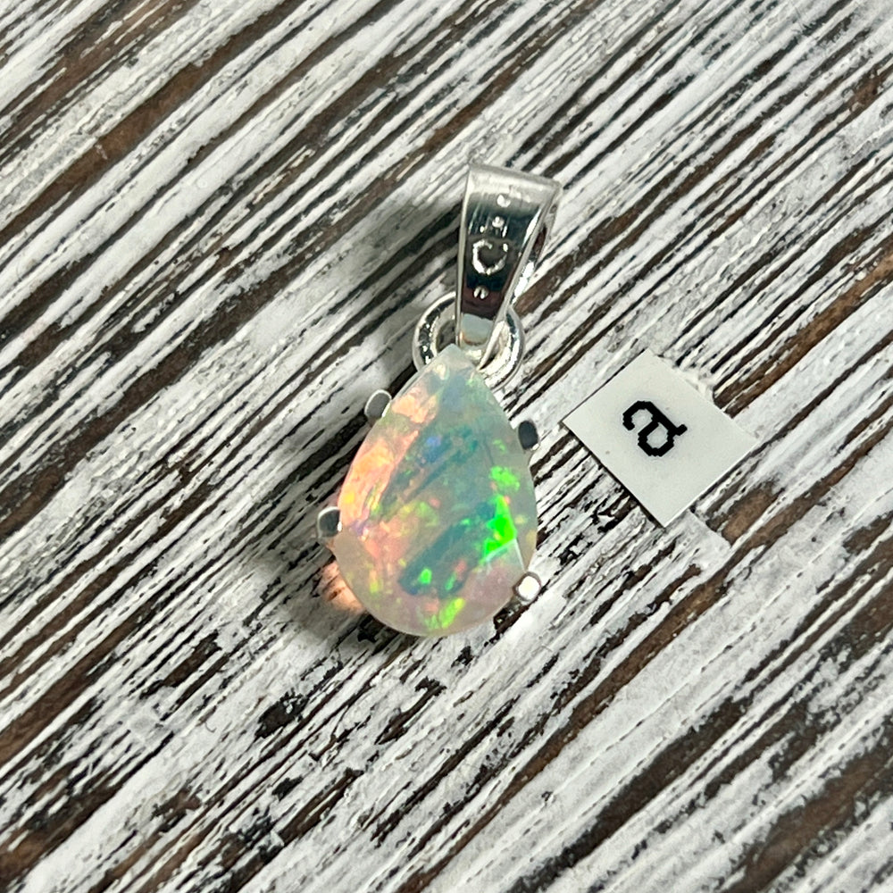 
                  
                    A Dainty Prong Set Facet Cut Teardrop Shaped Ethiopian Opal Pendant from Super Silver resting on a wooden table.
                  
                