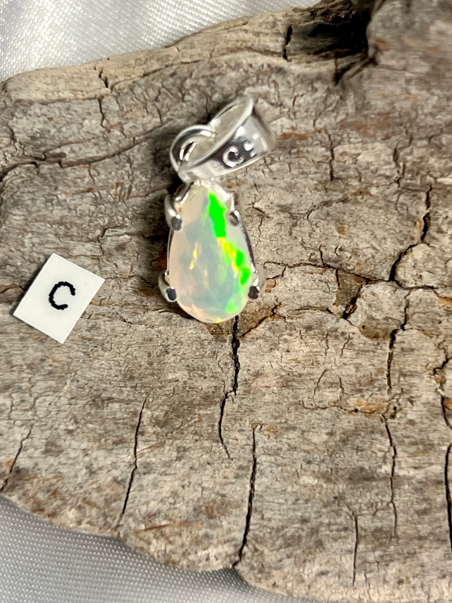 
                  
                    A Dainty Prong Set Facet Cut Teardrop Shaped Ethiopian Opal Pendant adorned with an exquisite Ethiopian Opal, gracefully suspended on a rustic piece of wood, by Super Silver.
                  
                