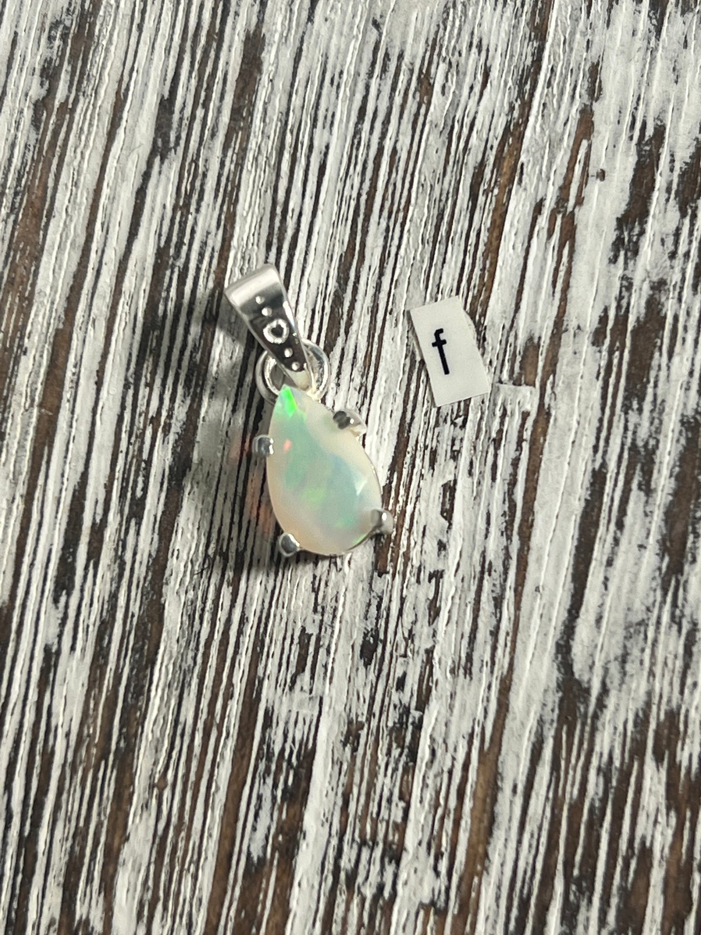 
                  
                    A Dainty Prong Set Facet Cut Teardrop Shaped Ethiopian Opal pendant by Super Silver on a wooden table.
                  
                