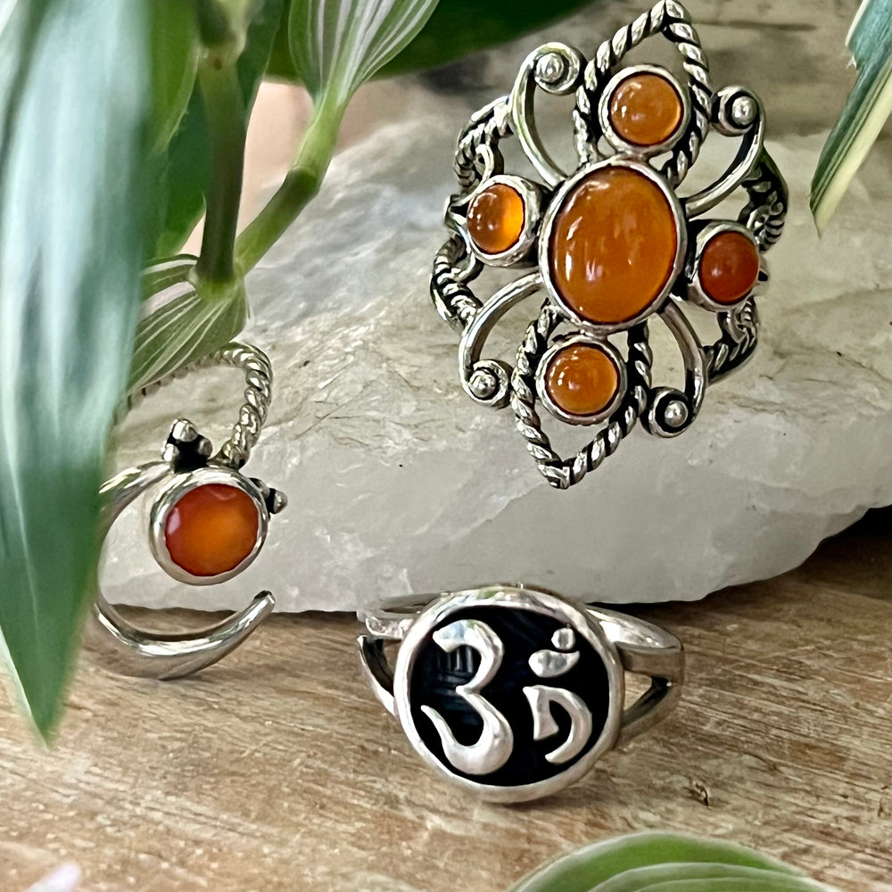 
                  
                    Three Online Only Exclusive Multistone Rings with carnelian gemstones and spiritual symbols displayed on a stone slab, surrounded by greenery.
                  
                