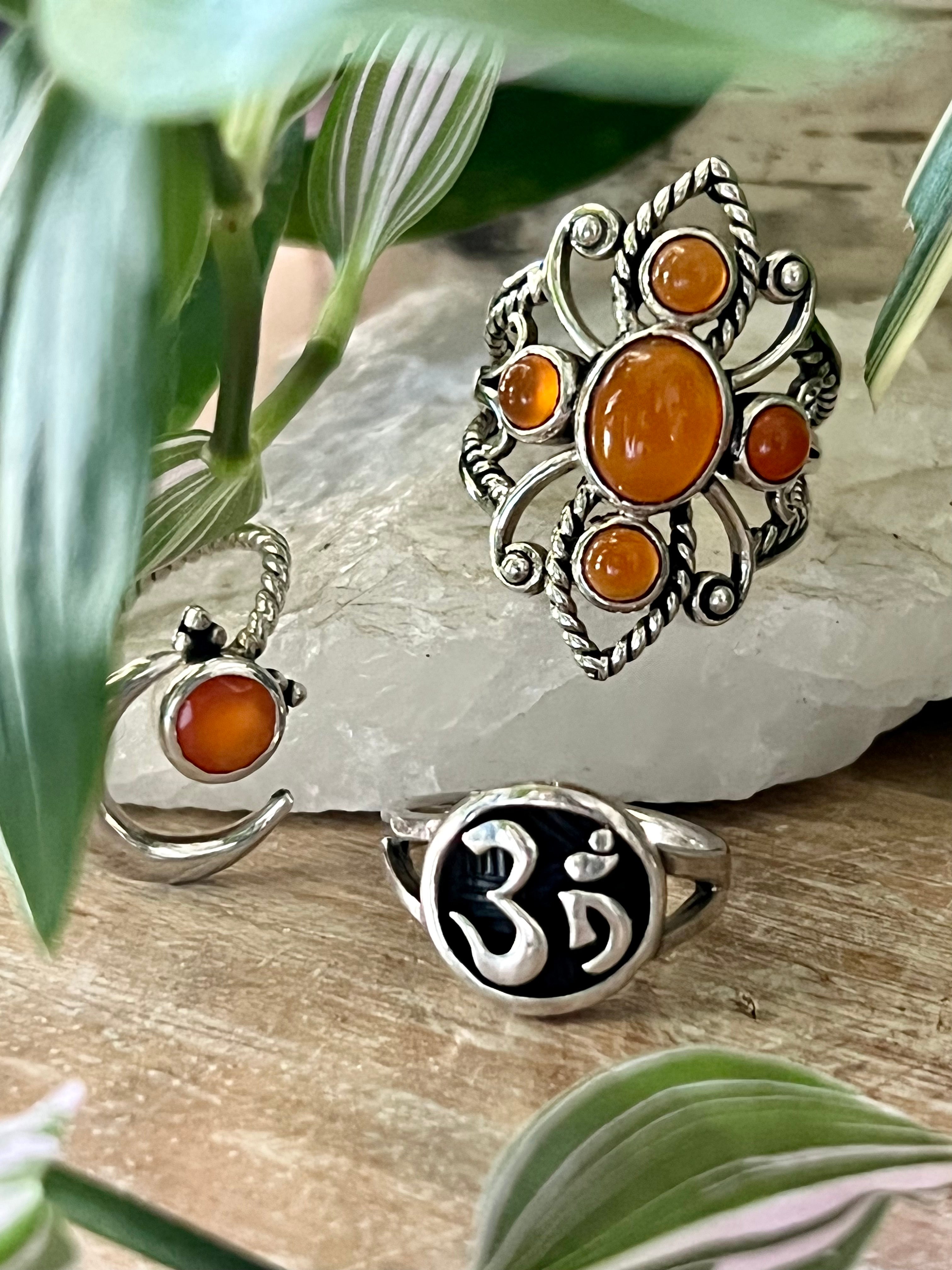 Three Online Only Exclusive Multistone Rings with carnelian gemstones and spiritual symbols displayed on a stone slab, surrounded by greenery.