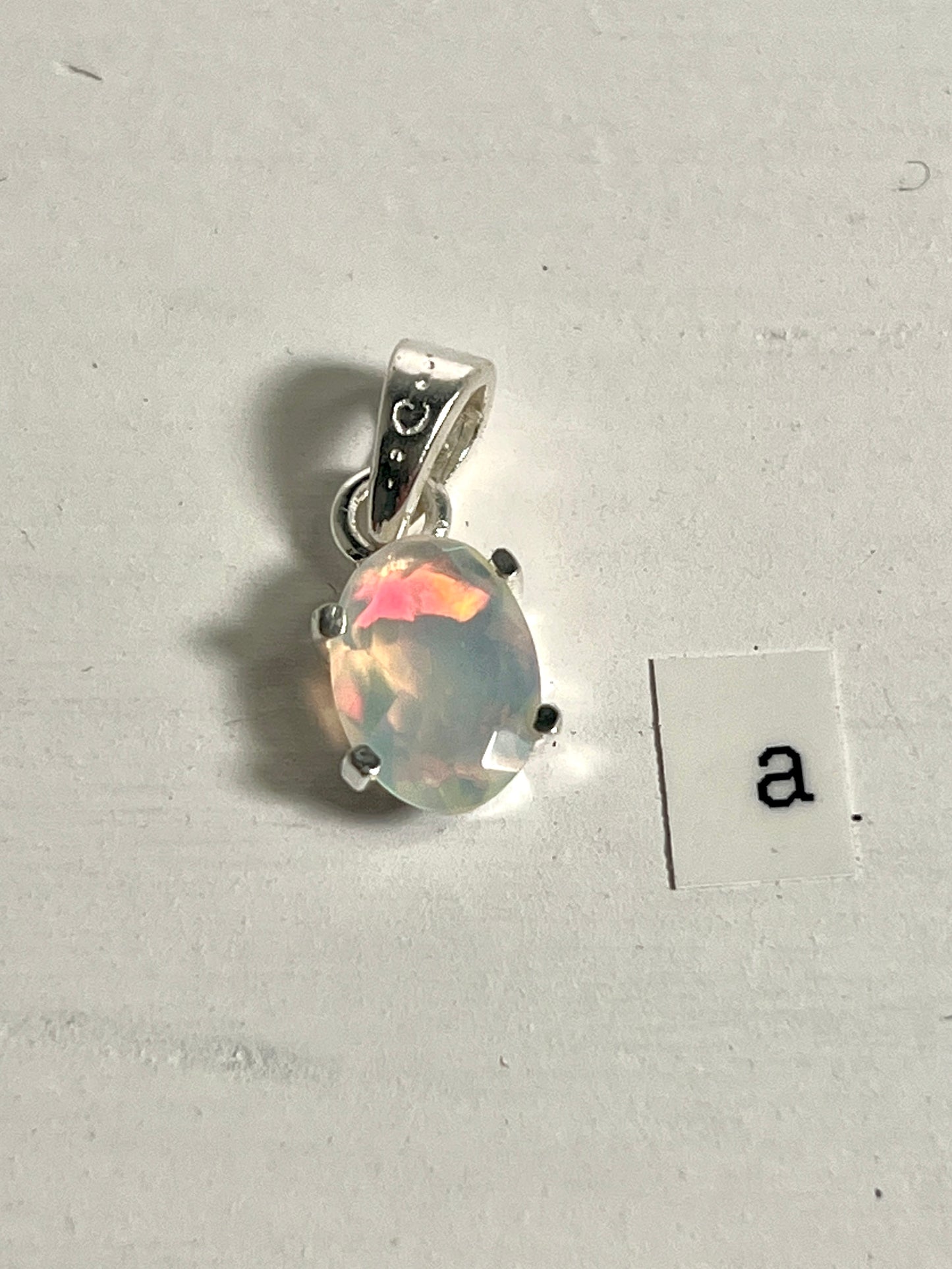 
                  
                    A tiny facet cut prong set Ethiopian Opal pendant with the letter a next to it, crafted in .925 sterling silver by Super Silver.
                  
                