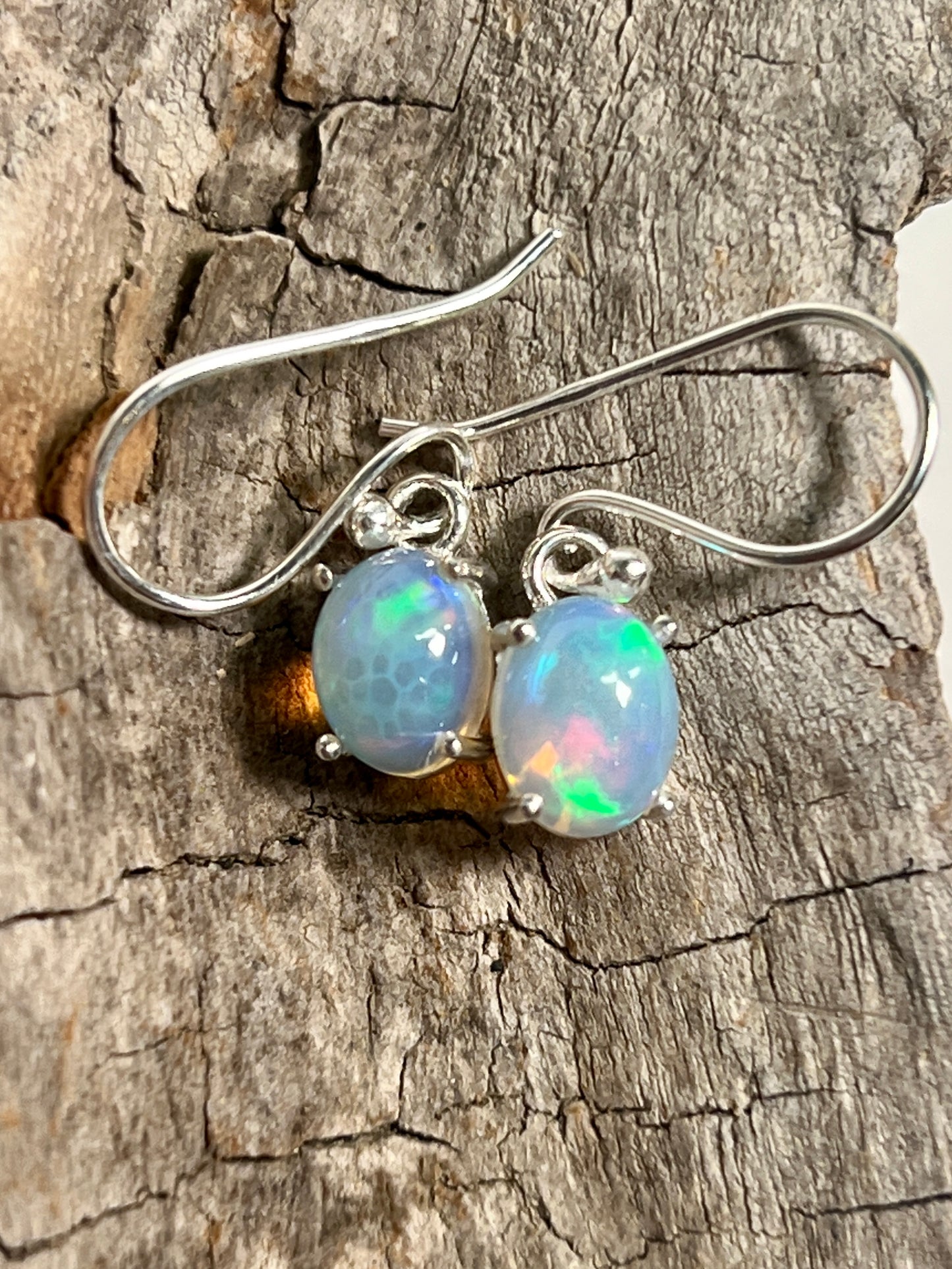 A pair of Vibrant Oval Ethiopian Opal Earrings from Super Silver, exuding natural brilliance on a piece of wood.