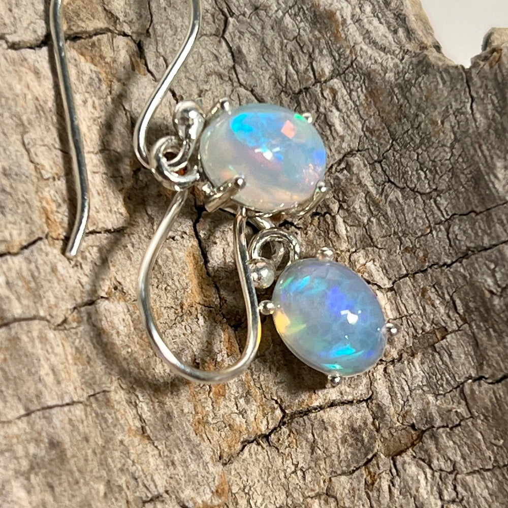 
                  
                    A pair of Super Silver Vibrant Oval Ethiopian Opal Earrings glowing with vibrant hues and natural brilliance, delicately displayed on a piece of wood.
                  
                