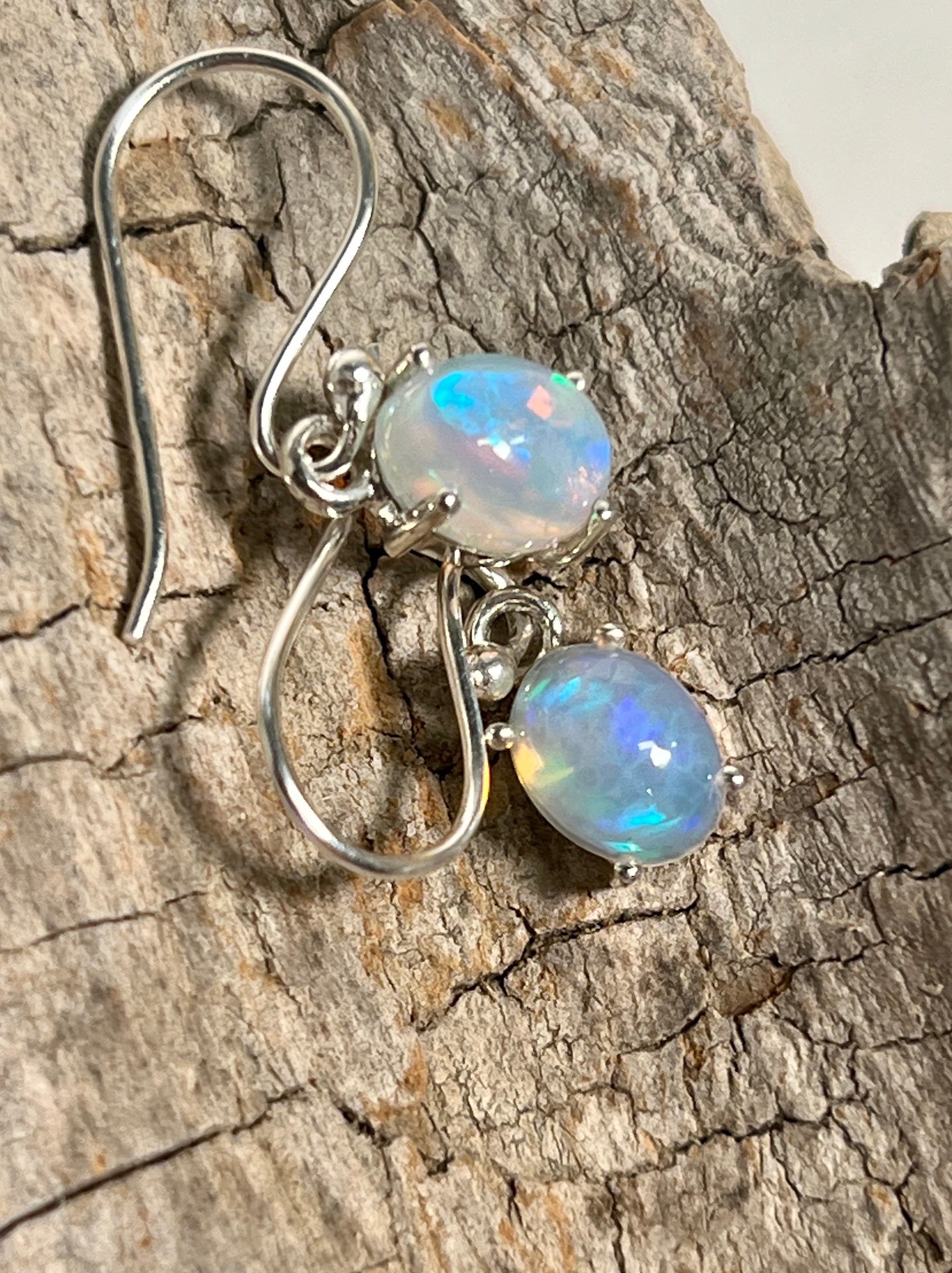 A pair of Super Silver Vibrant Oval Ethiopian Opal Earrings glowing with vibrant hues and natural brilliance, delicately displayed on a piece of wood.