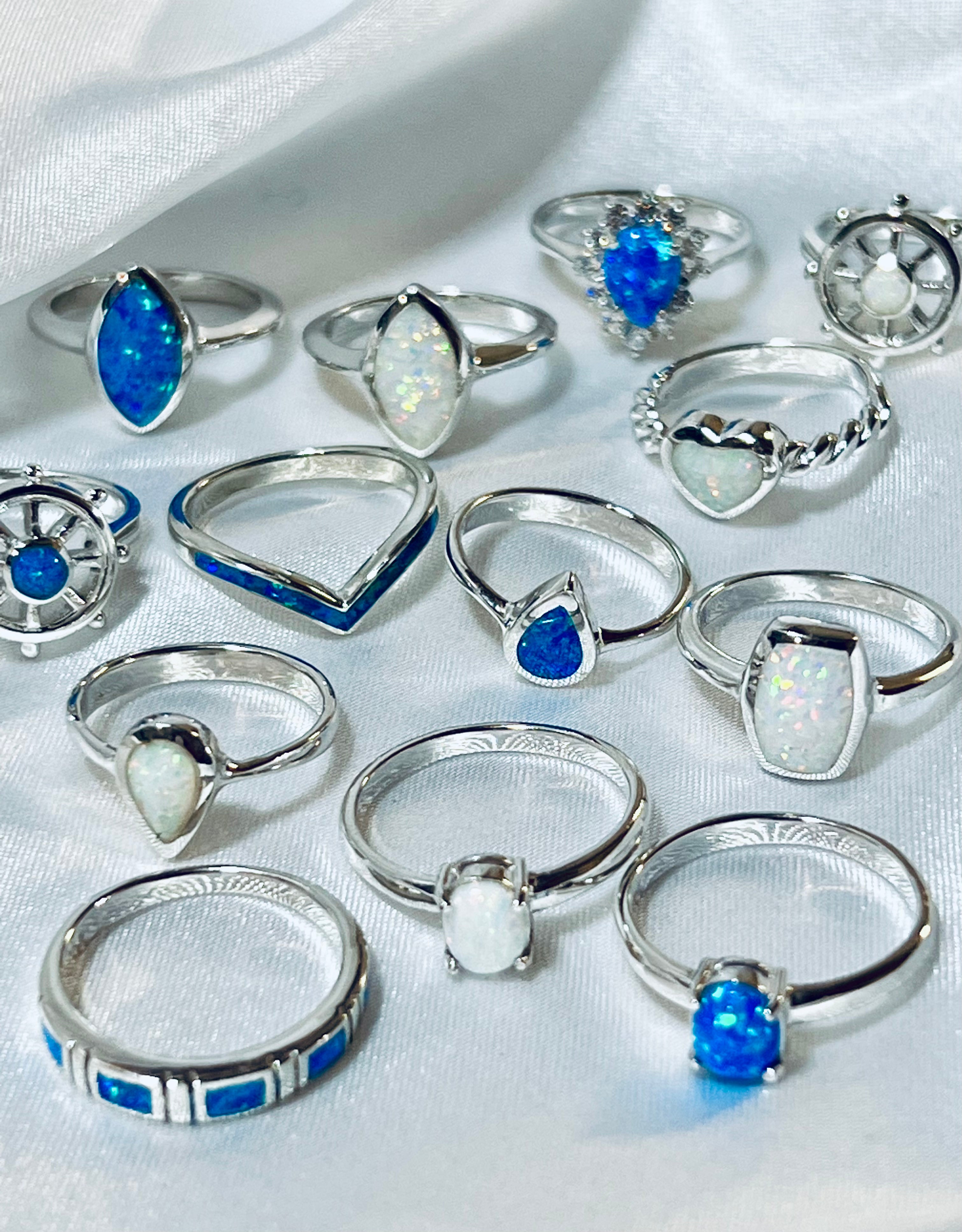 A collection of rhodium-plated silver rings with various blue and opalescent gemstones, including the Rectangular Lab Opal Ring, displayed on a white cloth.