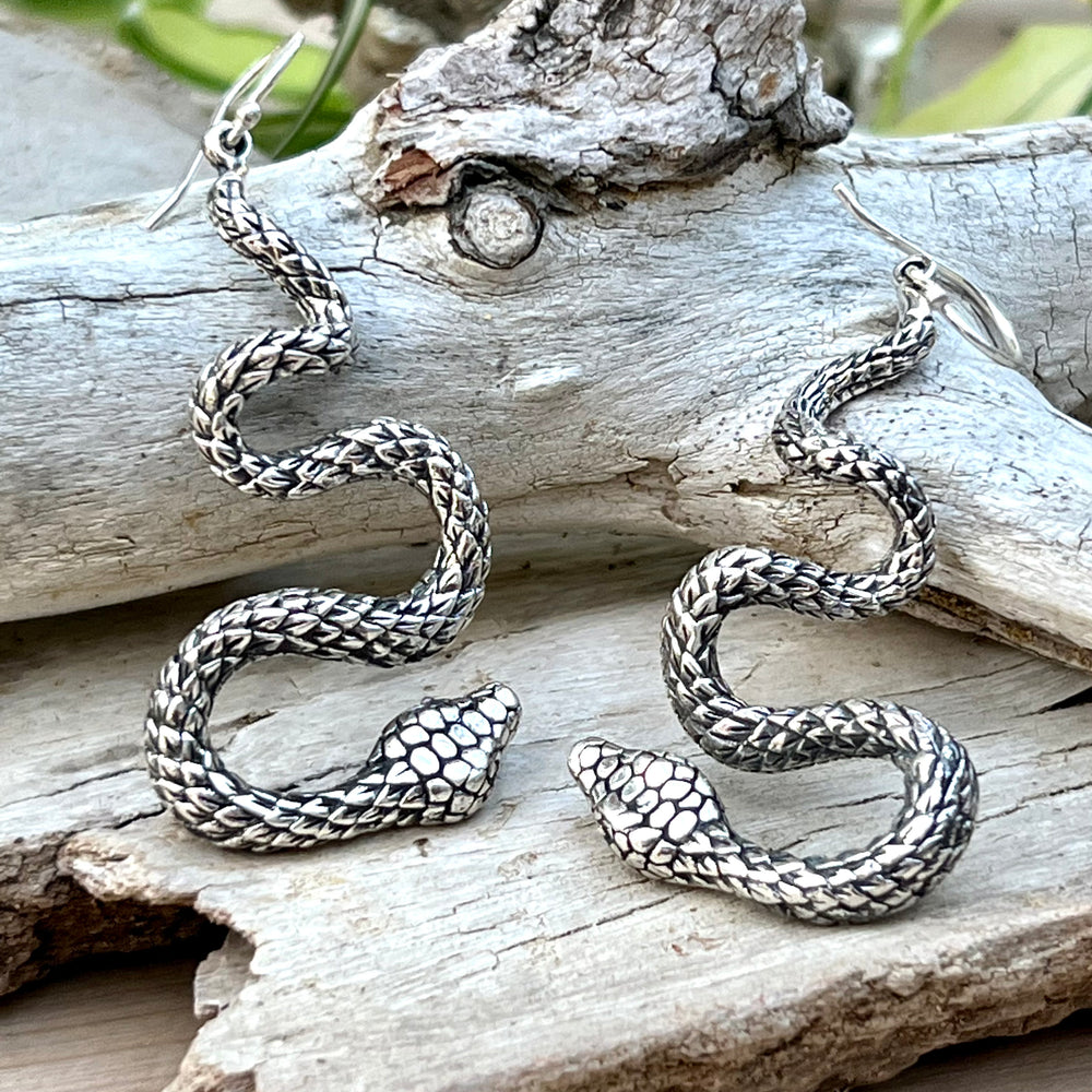 A pair of handcrafted Super Silver Long Twisting Snake Earrings on a piece of wood.