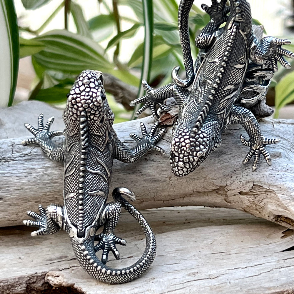 
                  
                    Two Handcrafted Super Silver Iguana Pendants sitting on a piece of wood.
                  
                