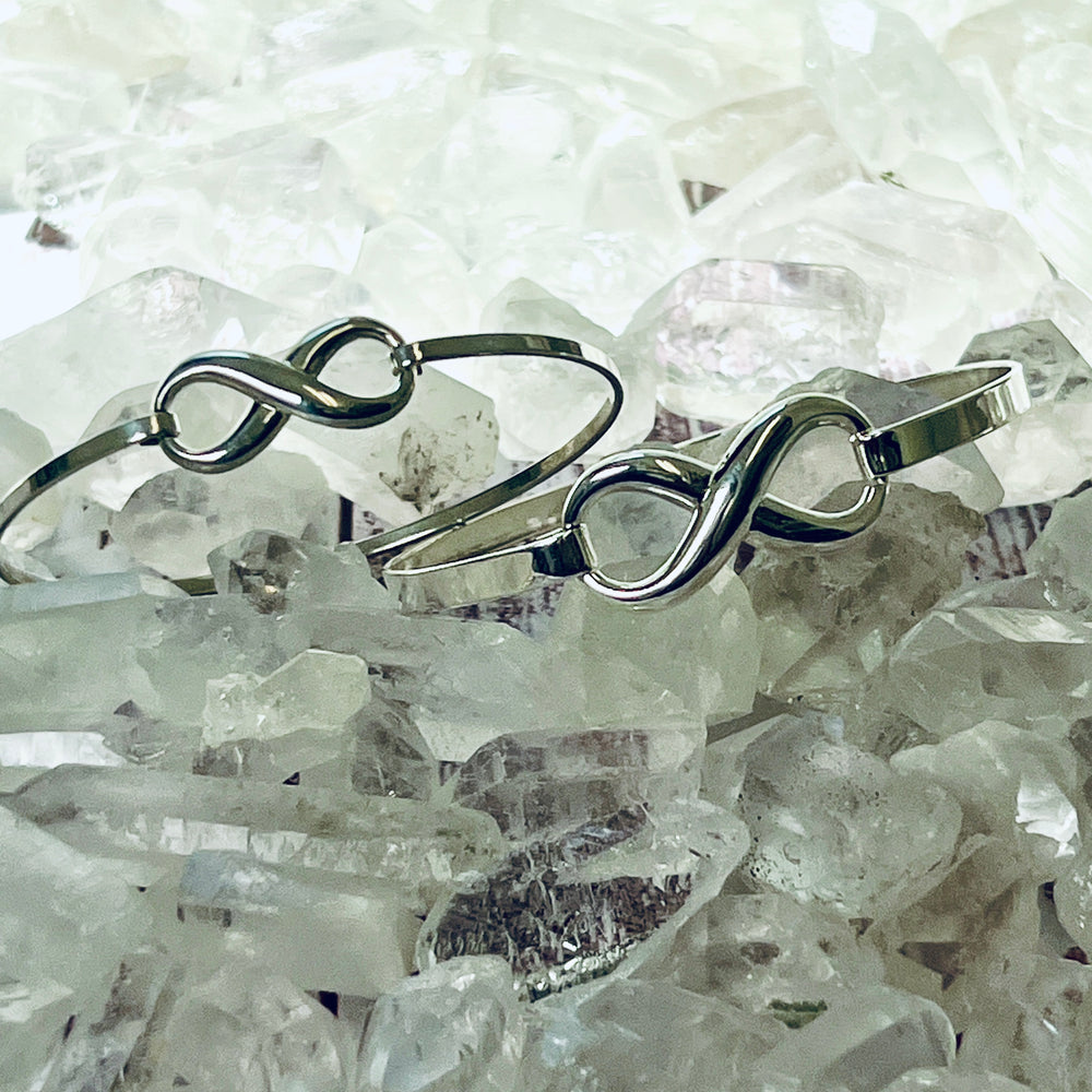 
                  
                    Two sleek silver Super Silver infinity bangles with a latch clasp on a pile of crystals.
                  
                