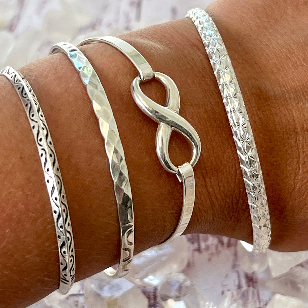 A woman's wrist adorned with three sleek Super Silver bracelets, featuring an elegant Infinity Bracelet and secured with a latch clasp.