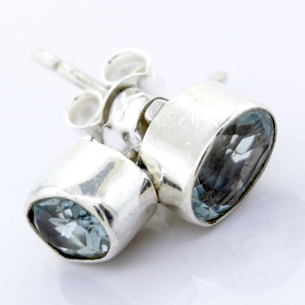 
                  
                    A pair of Beautiful Oval Faceted Cut Blue Topaz Studs by Super Silver with a silver setting on a white surface.
                  
                